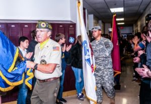 Veterans parade in school hallway with flags and uniforms.