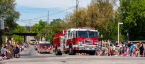 Fire trucks parade on sunny street with spectators watching.