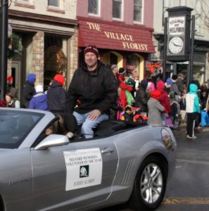 Man sitting on convertible in parade by florist shop.